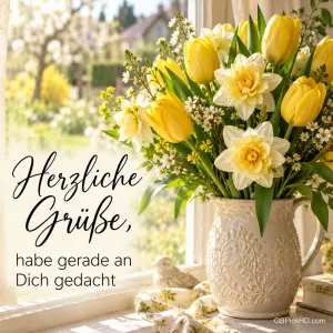 Weiße Keramikkanne mit gelben Tulpen und Narzissen auf einer sonnigen Fensterbank vor einem Frühlingsgarten, daneben der Text "Herzliche Grüße, habe gerade an Dich gedacht"