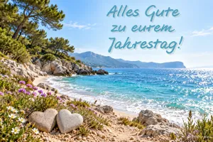 Zwei herzförmige Steine liegen am blühenden Meeresstrand unter "Alles Gute zu eurem Jahrestag!".