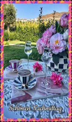 Gartentisch mit Tasse, Weinglas, rosa Blumen und grüner Gartenlandschaft am Nachmittag