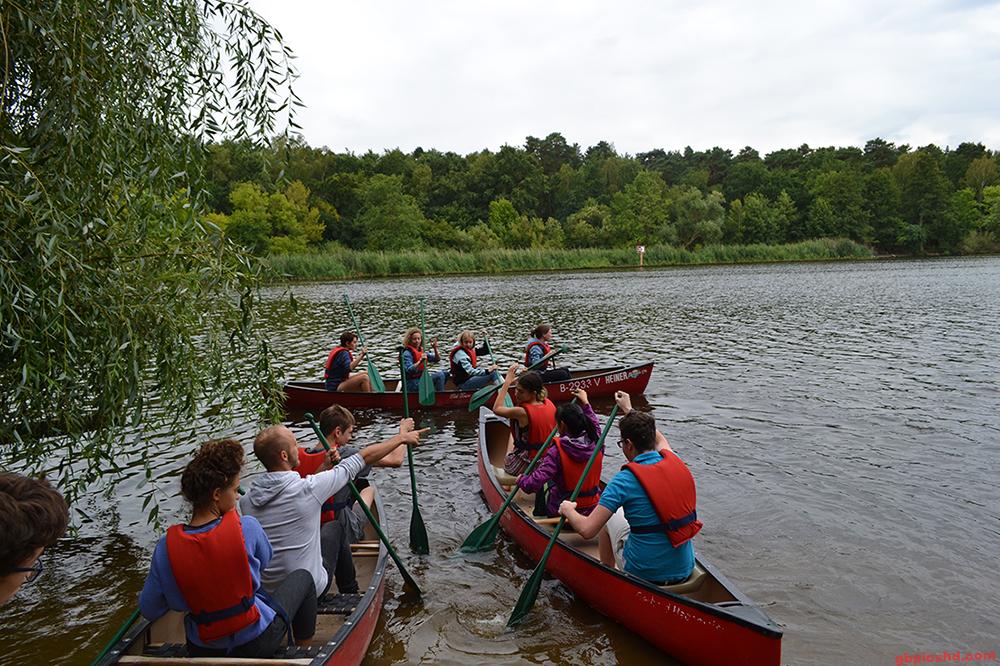 Menschen in Kanus auf einem See, umgeben von Bäumen, paddeln gemeinsam bei bewölktem Wetter.