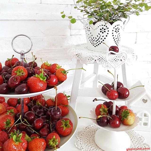 Zwei Etageren mit Erdbeeren und Kirschen vor weißem Hintergrund auf einem kleinen weißen Tisch mit Pflanze.